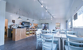 Modern café interior with wooden tables and chairs, counter with coffee machines and food display, hanging lights, and large windows creating a bright, inviting atmosphere.