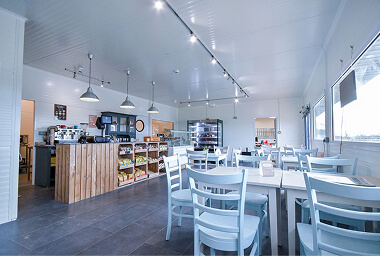 Bright café with white and light blue tables and chairs, wooden counter with coffee equipment and packaged goods, pendant lamps, and large windows creating an airy, welcoming atmosphere.