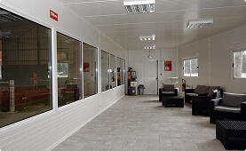 Clean lounge with wicker chairs and sofas with gray cushions, white paneled walls, tiled floor, and large windows, designed as a comfortable waiting or observation space.