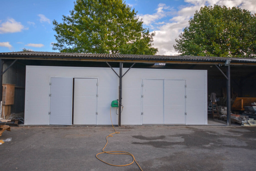 A metal storage shed with two closed roller doors and a side door, situated on a paved surface with trees in the background.