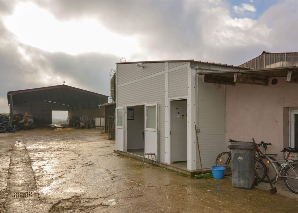 White modular building with bicycles, trash bin, and tools in foreground, large open barn with tractors and machinery in background, under cloudy sky with sunlight breaking through.