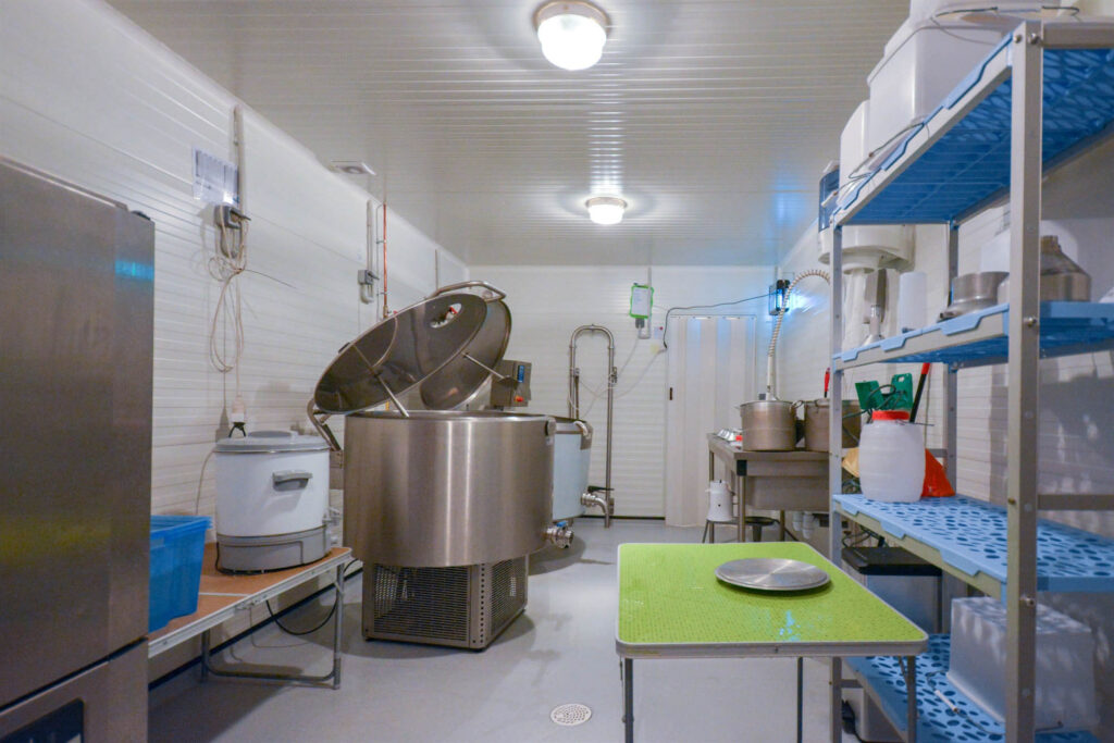 Clean food processing room with stainless steel mixing vat, yogurt or cheese maker, metal worktable, and blue shelving stocked with containers under bright overhead lighting.