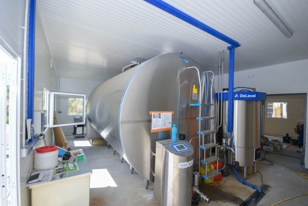 Stainless steel Packo milk cooling tank with pipes, valves, and ladder beside DeLaval tank and iControl unit in clean, well‑lit dairy processing room.
