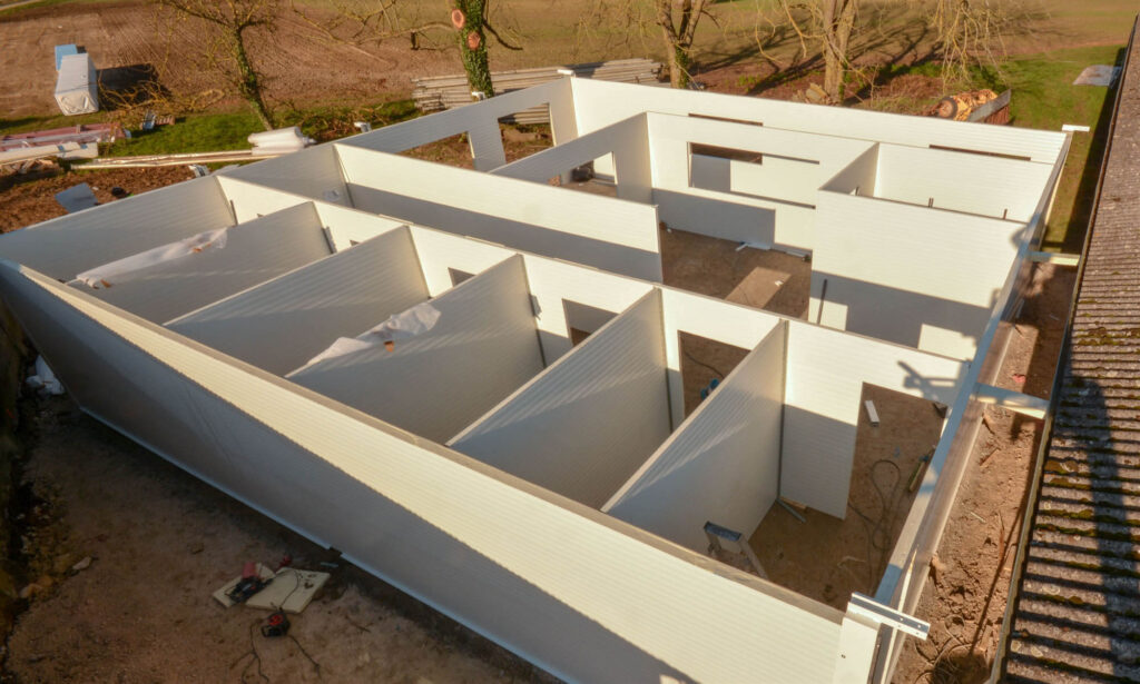 Partially assembled white modular building with prefabricated panels, multiple room layout visible, surrounded by construction tools, bare soil, and trees in background.