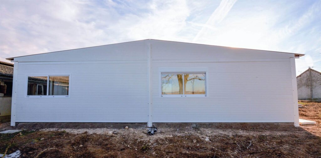 White modular building with gable roof and framed windows, set on bare soil with surrounding structures and trees in the background.